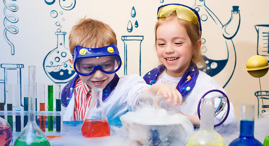 a boy and a girl wearing lab coats behind a desk with dry ice, beakers and tubes on it.